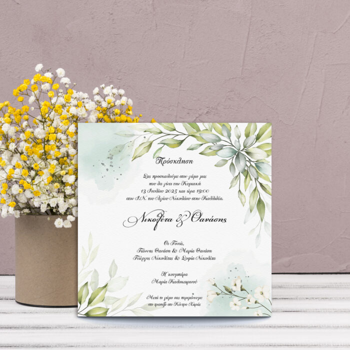 Πρόσκληση γάμου με πράσινα φύλλα και λευκά λουλούδια σε τετράγωνο σχήμα, μπροστά από βάζο με λουλούδια.