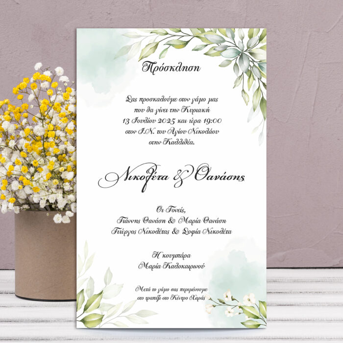 Πρόσκληση γάμου με πράσινα φύλλα και λευκά λουλούδια σε τετράγωνο σχήμα, μπροστά από βάζο με λουλούδια.