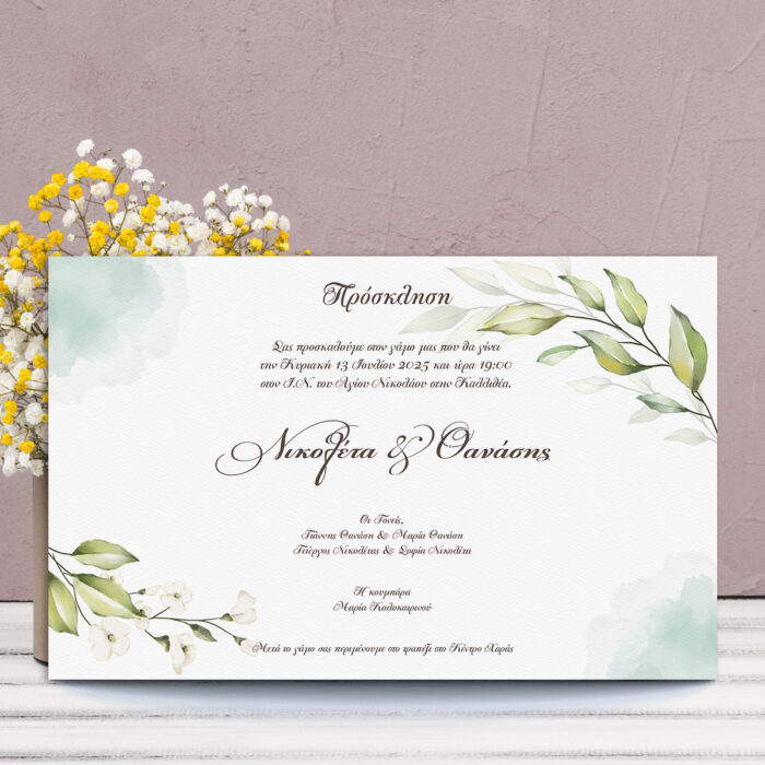 Πρόσκληση γάμου με πράσινα φύλλα και λευκά λουλούδια σε τετράγωνο σχήμα, μπροστά από βάζο με λουλούδια.